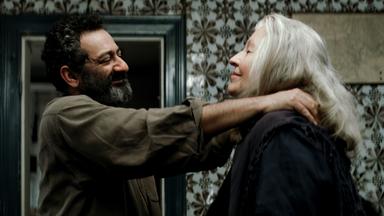 Ein Mann mit dunklen Haaren und graumeliertem Bart legt lächelnd einer älteren Frau mit langen grauen Haaren die Hände auf die Schultern.