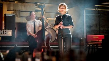 Monika (Sonja Gerhardt, r.) und Freddy (Trystan Pütter, l.) stehen auf einer Bühne. Monika hält ein Mikro in ihren Händen, während Freddy mit einem Cello neben ihr steht.