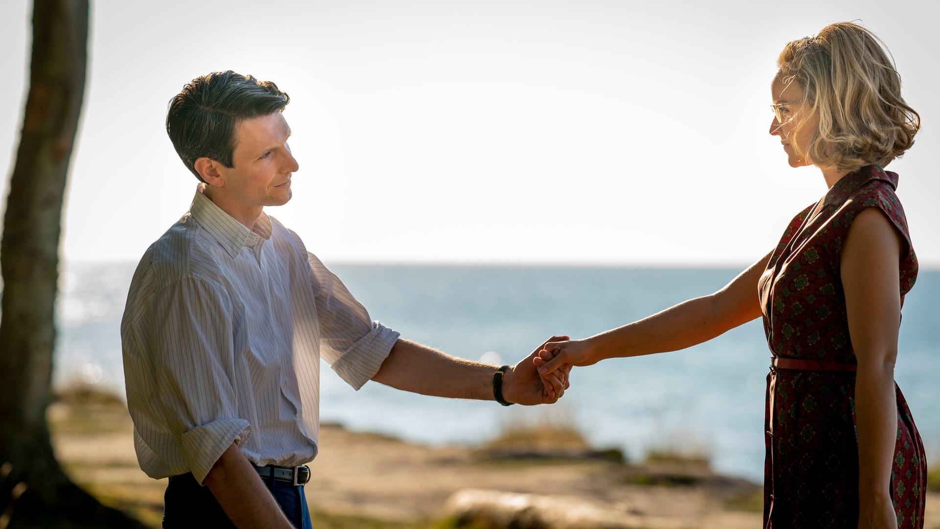 Joachim (Sabin Tambrea) und Monika (Sonja Gerhardt) genießen die Zeit am Meer. Sie stehen sich an einem Strand gegenüber und halten sich an einer Hand.