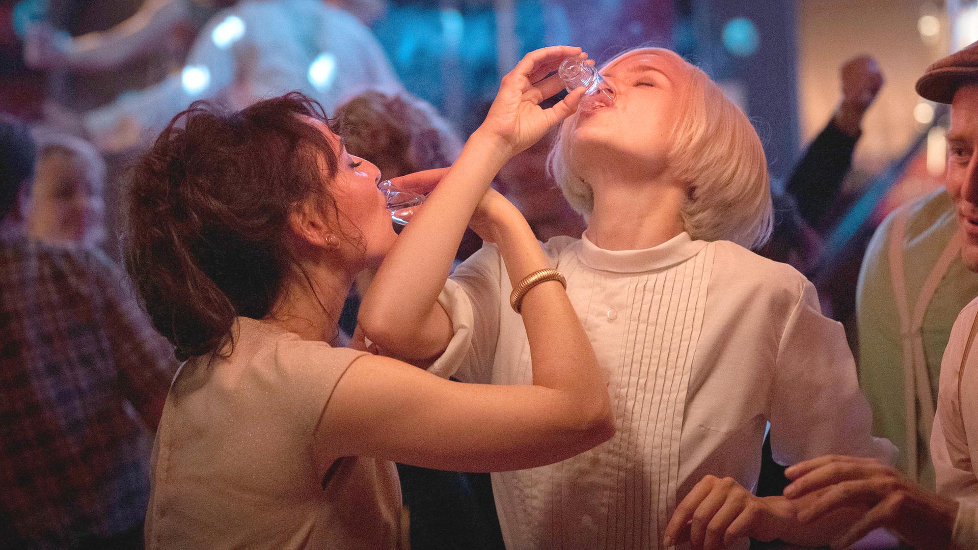 Helga (Maria Ehrich, l.) und Eva (Emila Schüle, r.) sind auf der Tanzfläche am Tanzen. Sie trinken dabei aus einem kleinen Glas Alkohol.