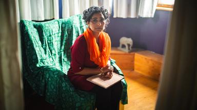 Szene aus "Meine Zuflucht und mein Sturm" von Arundhati Roy: Eine Fau sitzt auf einem grünen Sessel in einem Raum mit lila Wänden und weißen Vorhängen. Sie trägt ein rotes Oberteil und einen orangefarbenen Schal. Auf ihren Knien liegt ein Fotoalbum. Im Hintergrund steht eine kleine weiße Elefantenfigur auf einer Holzbank. Tageslicht fällt durch ein Fenster auf der rechten Seite ins Zimmer.