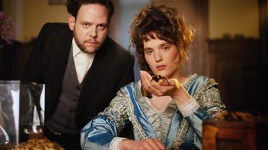 Szene aus "Die Unbehausten" von Barbara Kingsolver: Eine Frau in einem blauen Kleid sitzt an einem Holztisch. In ihrer linken Hand hält sie eine behaarte große Spinne. Daneben steht ein Mann im dunklen Anzug, der sich nach vorne beugt.