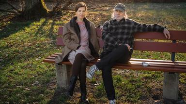 Nachgestellte Szene aus "Erzähl mir alles" von Elizabeth Strout: Eine Frau und ein Mann sitzen nebeneinander auf einer Holzbank in einem Park. Die Frau trägt einen hellen Mantel, ein Kleid mit schwarzen Stiefeln und sitzt mit übereinandergeschlagenen Beinen, während der Mann entspannt neben ihr sitzt und einen Arm auf der Rückenlehne der Bank ausstreckt. Er trägt eine karierte Jacke, dunkle Hose, Turnschuhe und eine Kappe. Neben ihm liegt eine Zigarettenschachtel auf der Bank. Im Hintergrund Bäume und winterliches Gras im warmen Sonnenlicht.