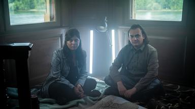 Szene aus "Was wir wissen können" von Ian McEwan: Zwei Personen sitzen auf dem Holzboden eines Zimmers mit zwei Fenstern, durch die man grüne Landschaft und Wasser sieht. Sie sitzen auf Decken, vor ihnen liegt eine große Landkarte ausgebreitet. Hinter ihnen stehen eine Lampe und zwei vertikale Leuchten, die die Szene beleuchten. Die Atmosphäre wirkt ruhig und konzentriert.