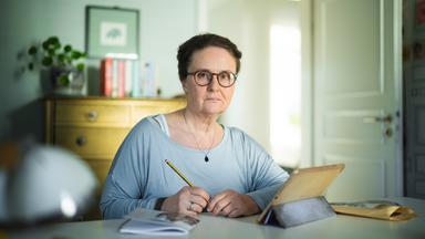 Nachgestellte Szene aus "Ich möchte zurückgehen in der Zeit" von Judith Hermann: Eine Frau sitzt an einem Tisch und hält einen Bleistift in der Hand. Sie trägt eine Brille und blickt mit ernstem Ausdruck nach vorne. Vor ihr stehen ein Tablet in einer Hülle, daneben liegt ein Notizbuch und ein brauner Briefumschlag. Hinter ihr befindet sich ein heller Raum mit einer Kommode, Büchern und einer Zimmerpflanze, durch eine offene Tür fällt Tageslicht hinein.