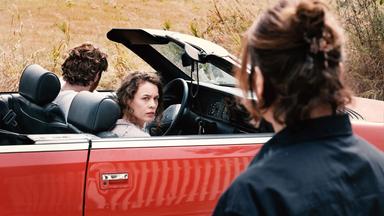 Eine Frau schaut auf ein rotes Cabrio, in dem ein Mann und eine Frau sitzen. Der Mann sitzt hinterm Steuer, die Frau schaut vom Beifahrersitz auf die betrachtende Frau. Von dieser ist nur der Hinterkopf zu sehen.