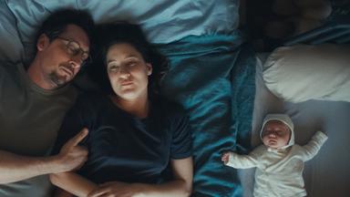 Ein Mann und eine Frau liegen auf ihrer Bettdecke im Bett. Der Mann hat die Augen geschlossen und lehnt seinen Kopf an den der Frau. Auch mit seinem Arm hält er sich an ihr fest. Die Frau hat die Augen geöffnet und blickt ins Leere. Neben ihnen im Beistellbett liegt ein schlafendes Baby.
