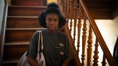 Szene aus "Der Fluch der Falodun Frauen" von Oyinkan Braithwaite: Eine junge Frau steht auf einer Holztreppe mit kunstvoll geschnitztem Geländer und blickt direkt in die Kamera. Sie trägt ein graues T-Shirt mit mehreren Aufnähern und hat eine helle Tasche über der Schulter. Im Hintergrund befindet sich eine rote Tür.