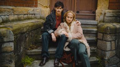 Inszenierte Szene zum Buch "Meine Liebe stirbt nicht" von Roberto Saviano: Vor dem Eingang eines Gebäudes mit Backsteinmauer und großer Holztür sitzt ein junges Paar auf einer steinernen Treppe. Der Mann trägt eine dunkle Jeansjacke und hat seinen Arm um die Frau gelegt, die einen hellen Mantel mit Fellbesatz und ein gemustertes Stirnband trägt. Beide schauen ernst in Richtung Kamera.