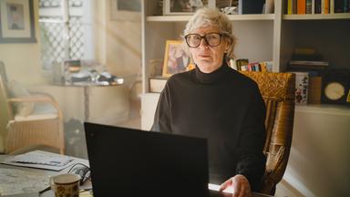 Nachgestellte Szene aus "Ghost Stories. Ein Buch der Erinnerung" von Siri Hustvedt: Eine ältere Frau mit kurzen grauen Haaren und runder Brille sitzt in einem Korbstuhl vor einem Laptop. Sie trägt einen schwarzen Rollkragenpullover. Vor ihr stehen eine Tasse und mehrere Briefe, im Hintergrund sind Bücherregale, Familienfotos und eine Uhr zu sehen. Durch ein Fenster fällt warmes Tageslicht in den Raum und erzeugt eine ruhige Atmosphäre.