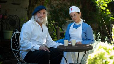 Szene aus  "Herz in der Faust" von Sorj Chalandon: Ein älterer Mann und eine Frau sitzen an einem kleinen runden Tisch im Freien. Der Mann trägt ein weißes Hemd, schwarze Hose und eine blaue Baskenmütze. Die Frau ist in einer Krankenschwesteruniform mit einem blauen Kleid, weißer Schürze und einer Haube mit rotem Kreuz gekleidet. Beide haben eine Tasse vor sich, im Hintergrund ist ein schattiger Garten mit Pflanzen und Bäumen zu sehen.