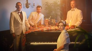 Szene aus "Das Haus der Türen" von Twan Eng Tan: Eine Frau sitzt vor der Tastur eines Flügels und blickt in die Kamera. Um den Flügel stehen drei Männer in weißen Anzügen. Der Flügel ist bedeckt mit einer rot gemusterten Tischdecke, einem Sektkühler mit Flasche und verschiedenen Gläsern.