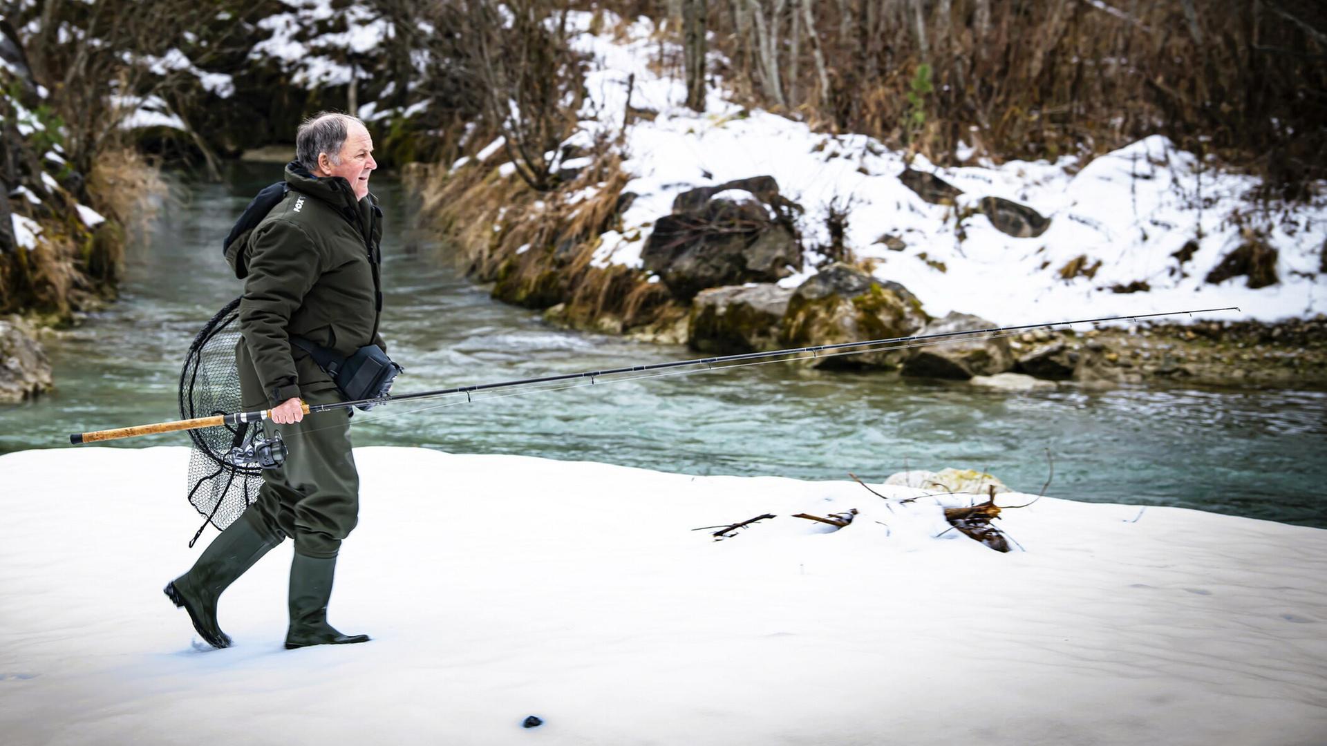 ein Angler in Montur stapft durch das verschneite Ufergelände auf den Bach zu