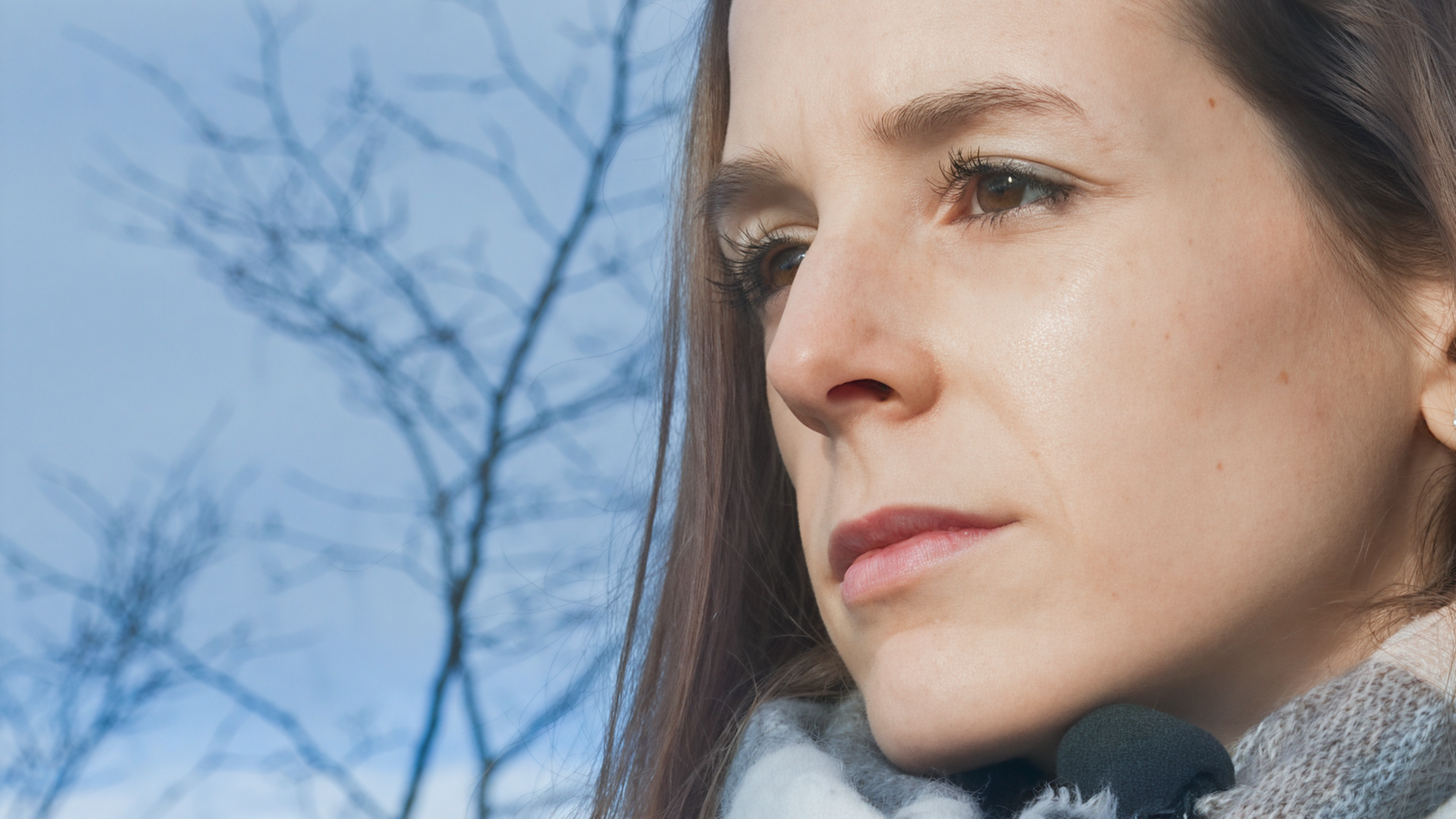 Eine Frau mit langen braunen Haaren schaut ernst in die Ferne; im Hintergrund kahle Äste und ein blauer Himmel.