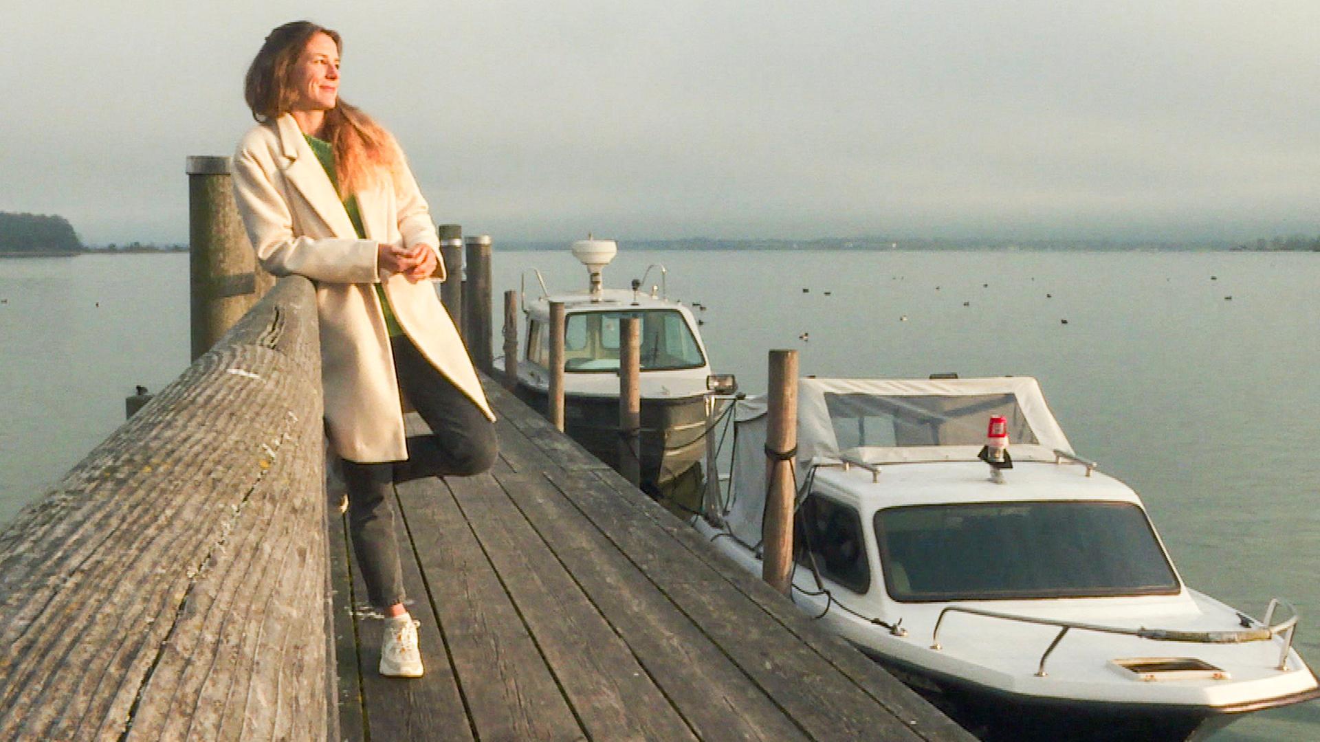 Eine Frau lehnt lächelnd an einem Geländer auf einem Bootssteg, im Hintergrund ein See mit Motorbooten