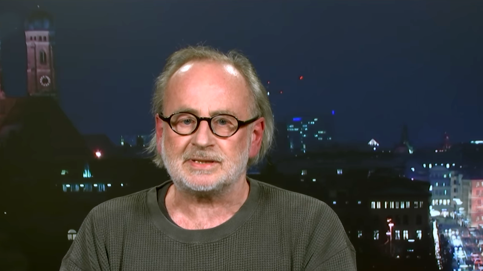 Porträtfoto eines Mannes mit Glatze und Brille vor dem Hintergrund einer beleuchteten Stadt bei Nacht.