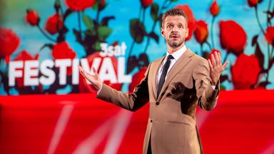 Kabarettist steht in der Mitte des Bildes und breitet seine Arme seitlich aus. Die Bühne ist rot beleuchtet und es sind Rosen auf der Vidiwall abgebildet. Florian Schroeder trägt einen beigen Anzug.