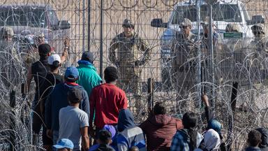 Rückansicht von Menschen dunklerer Hautfarbe, die vor einem Stacheldrahtzaun stehen. Auf der anderen Seite stehen Soldaten in Camouflage.