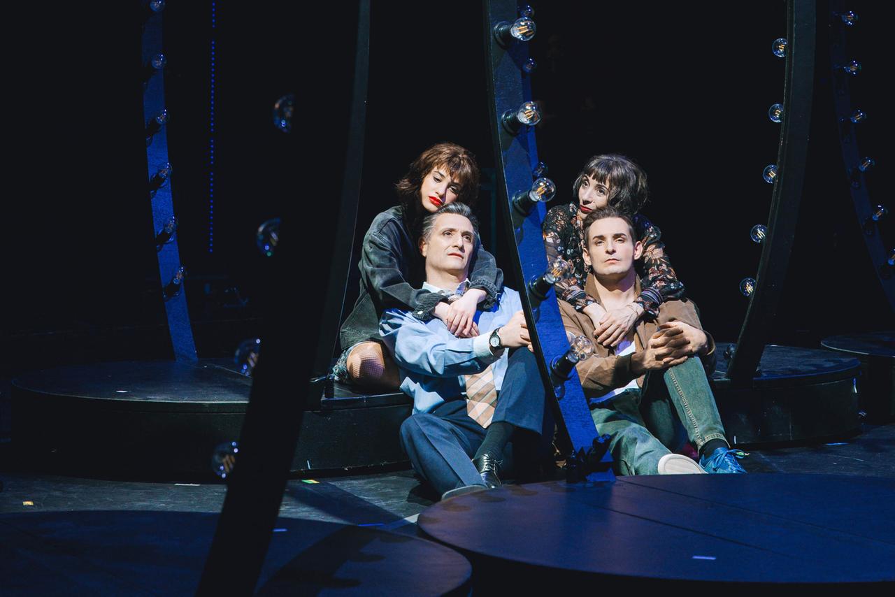 Das Bild zeigt eine Szene aus dem Musical "Follies". Im Mittelpunkt sind vier Personen, die auf einer dunklen Bühne sitzen.   Die Bühne ist mit kreisförmigen Podesten ausgestattet, und es gibt mehrere große, runde Spiegelrahmen, die von Lampen umgeben sind und Licht reflektieren. Diese Lichter sind ausgeschaltet und erzeugen eine stimmungsvolle, dunkle Atmosphäre.  Die Personen sind wie folgt angeordnet:   1. **Vorne links**: Eine Frau mit lockigen, dunklen Haaren und einem schwarzen Kleid, die sich an einen Mann anlehnt. 2. **Vorne Mitte**: Ein Mann mit grauen Haaren, der in einem blauen Hemd und einer gestreiften Krawatte gekleidet ist, wirkt nachdenklich, während er die Arme um die Oberschenkel gelegt hat. 3. **Hinten rechts**: Eine weitere Frau mit dunklen, kurzen Haaren und einem floralen Oberteil, die sich an den hinteren Mann lehnt. 4. **Hinten links**: Ein junger Mann mit kurzen, etwas unordentlichen Haaren, der in einem braunen Oberteil und einer Jeans sitzt.  Die gesamte Szene vermittelt eine melancholische Stimmung, die sowohl Nostalgie als auch Verbundenheit zwischen den Charakteren ausdrückt.