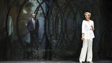 Das Bild zeigt eine Szene aus dem Musical "Follies", das an der Wiener Volksoper aufgeführt wird. Im Vordergrund stehen zwei Figuren auf der Bühne.   Die Frau trägt einen weißen, eleganten Hosenanzug mit einem taillierten Schnitt und hat eine goldene Tasche in der Hand. Ihr kurzes, gewelltes Haar ist hellblond. Sie hat einen nachdenklichen Ausdruck und schaut leicht zur Seite.  Im Hintergrund steht ein Mann, der in einem dunklen Anzug mit einer gestreiften Krawatte gekleidet ist. Er hat die Hände in die Taschen gesteckt und blickt ernst auf eine transparente, strukturelle Kulisse. Diese Kulisse zeigt mehrere ringförmige Lichtinstallationen, die teilweise beleuchtet sind und einen nostalgischen Eindruck vermitteln.  Der Gesamteindruck des Bildes strahlt eine gewisse Melancholie und Dramatik aus, passend zur Thematik des Musicals, das sich mit Erinnerungen und der Vergänglichkeit von Träumen befasst.