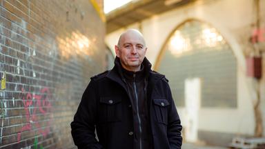 Der Schriftsteller Frank Goosen steht vor einer Ziegelmauer, im Hintergrund sind ein Torbogen und Paletten zu sehen.