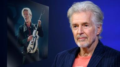 Ein älterer Mann mit grauem Haar und in T-Shirt und Jacket blickt mit einem dezenten Lächeln in die Kamera. Links von ihm ist ein Bild von einem Mann, der auf der Bühne E-Gitarre spielt, eingeblendet