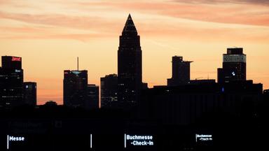 Der Messeturm und weitere Hochhäuser in Frankfurt dunkel vor einem gelb-roten Sonnenuntergang.