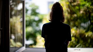 Rückansicht einer jungen Frau mit zusammengebundenen Haaren, die aus einem geöffneten Fenster schaut.