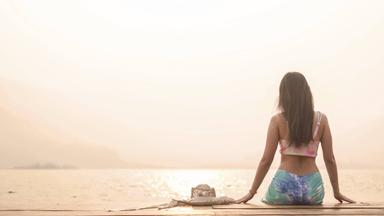 Eine Frau in Sport-Top und Leggings sitzt mit dem Rücken zum Betrachter auf einem Steg am Meer oder an einem großen See.