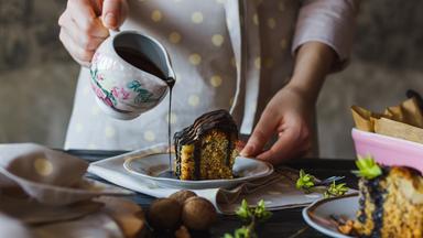 Eine Frau gießt Schokoladensauce über ein Stück Kuchen, das auf einem Dessertteller angerichtet ist.