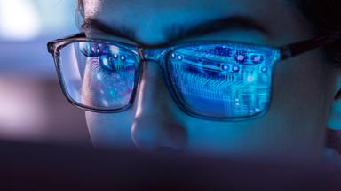 Frau sitzt hinter Computer. Virtuelle Daten leuchten auf ihren Brillengläsern