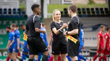 Fabienne ist im Stadion in Schiedsrichteroutfit im Gespräch mit zwei männlichen Kollegen. Im Hintergrund laufen Fußballspieler in blauer oder roter Trainingskleidung.