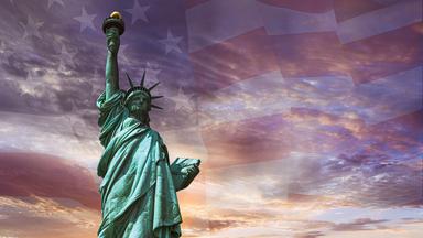 Die Freiheitsstatue - eine Frau mit Sternenkrone hält eine Fackel hoch - vor verschwommener US-Flagge