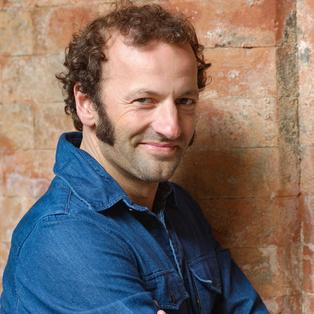 Der Moderator im Jeanshemd vor einer Wand