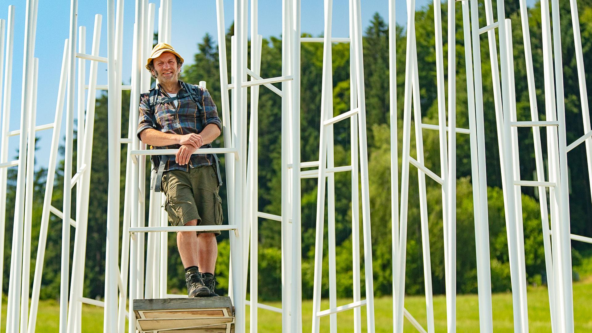 Der Schmid Max steht erhöht zwischen unterschiedlich angeordneten, weißen Leitern – ein Kunstwerk des Architekten Sou Fujimoto.