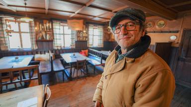 Schmidt Max im Gastraum eines Gasthofs. Dort stehen einfache Tische und Stühle aus Holz vor Sprossenfenstern mit karierten Vorhängen.