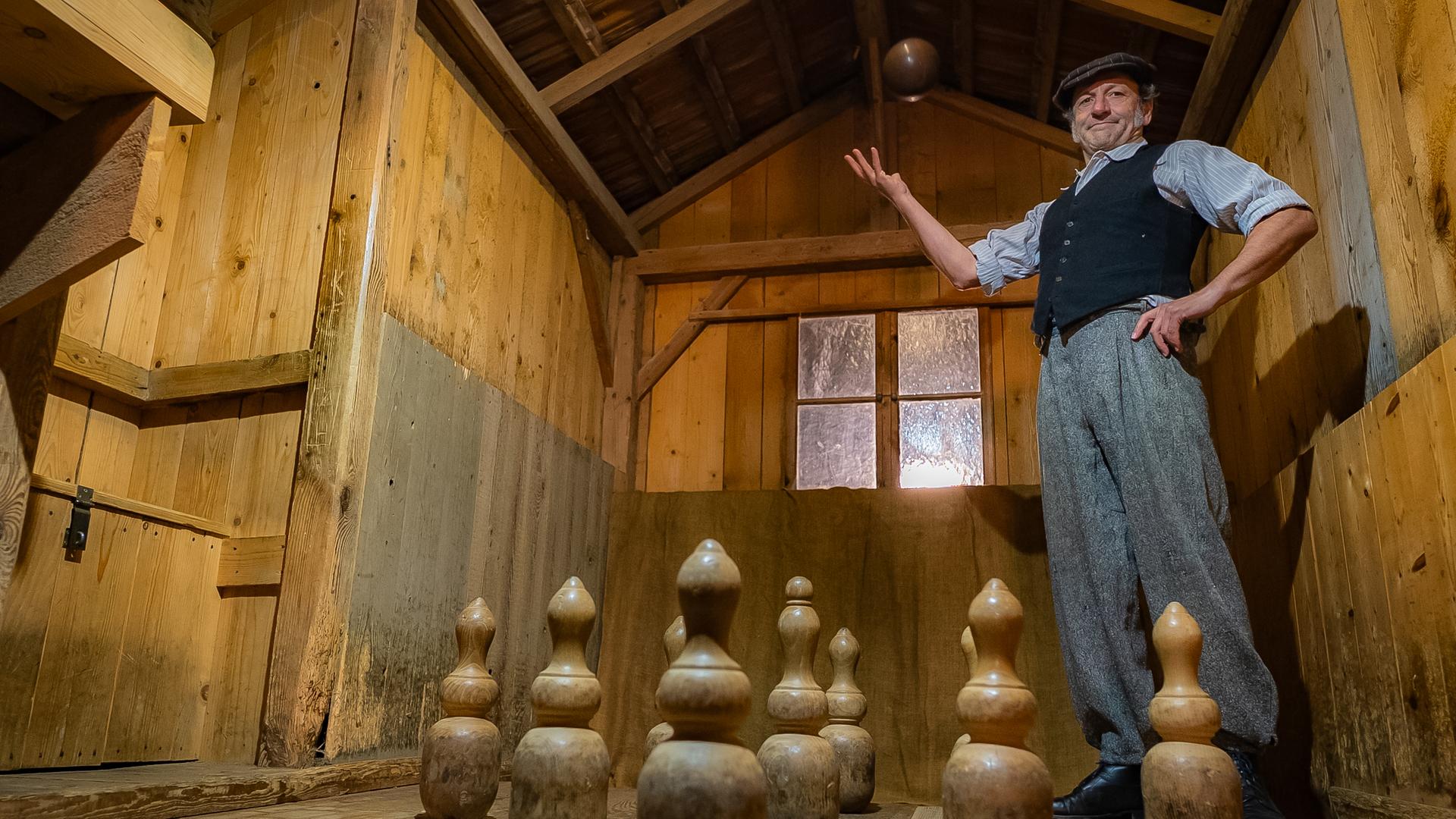 Schmidt Max  steht in einer historischen Kegelbahn, in der alles aus Holz ist: Die Kegel, die Kugel, Wände und Kegelbahn.