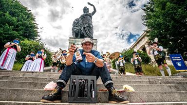 Schmidt Max sitzt unterhalb der Bavaria auf einer Treppe mit einer Flasche in der Hand daneben steht eine Trachtlergruppe