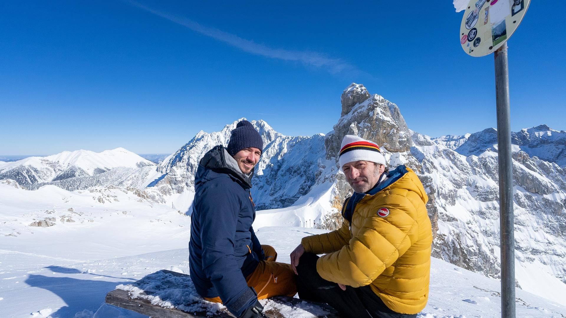 Zwei Männer sitzen auf einer Bank im Schnee vor Bergkulisse. 