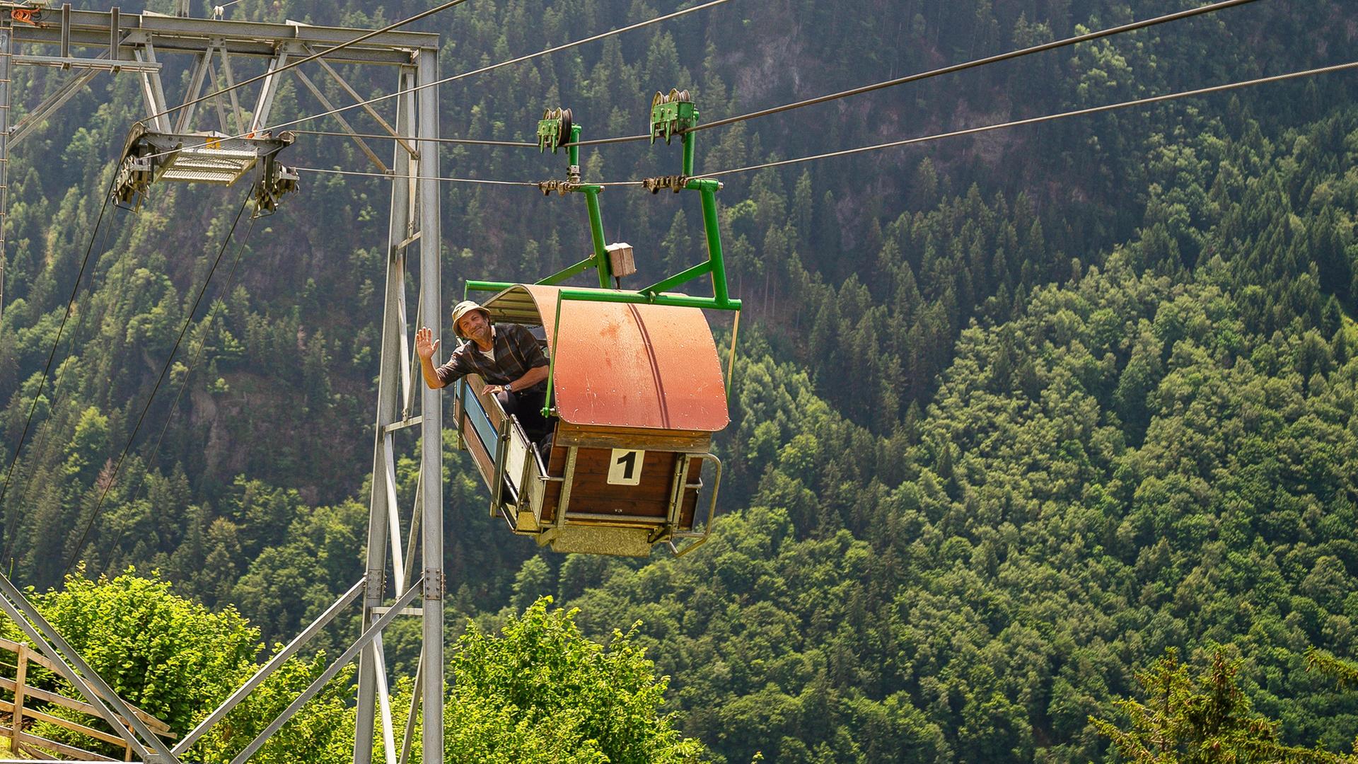 Schmidt Max winkt aus der Gondel einer Seilbahn.