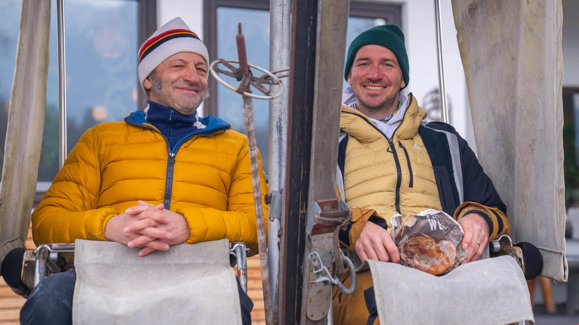Schmidt Max und der ehemalige Skirennfahrer Felix Neureuther sitzen lächelnd in den stillstehenden alten Sitzen einer Sesselbahn.