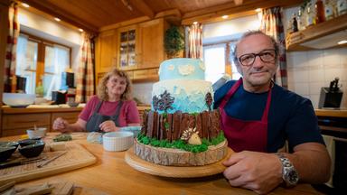 Konditormeisterin Rosi Sigl und Schmidt Max sitzen an einem Tisch. Darauf steht eine dreistöckige Torte, die mit einem Igel und einer Landschaft verziert ist.