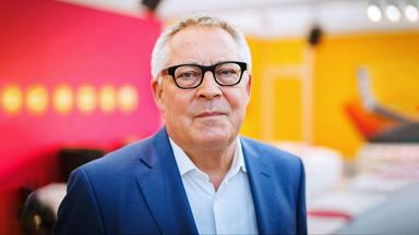 Porträt Karl Schlögel: Der Historiker blickt in die Kamera, trägt eine Brille und einen dunklen Blazer. Der Hintergrund ist neutral in den Farben orange und rot.