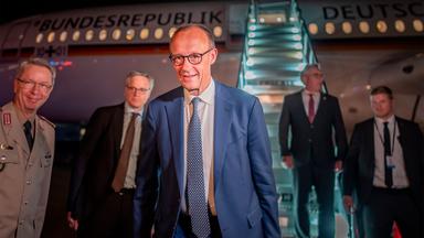 Mehrere Männer in Anzügen stehen in der Dämmerung vor einem Flugzeug, an das eine Rolltreppe angedockt ist.