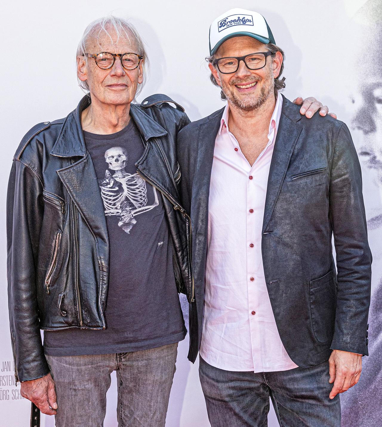 Rainer Pause und Aljoscha Pause stehen nebeneinander und lächeln in die Kamera. Rainer Pause hat seinen Arm um Aljoscha Pauses Schulter gelegt. Rainer Pause trägt eine schwarze Motorradlederjacke und darunter ein schwarzes Shirt mit der Aufdruck eines menschlichen Skeletts. Aljoscha Pause trägt ein schwarzes Ledersakko und eine Baseball-Kappe. Sie stehen neben dem Filmplakat mit ihren Konterfeis.