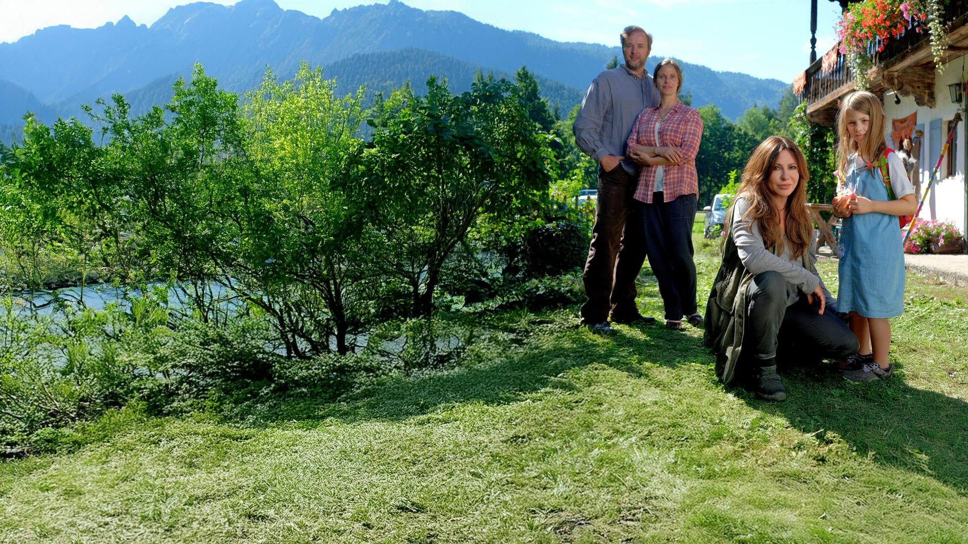 "Frühling - Wenn Kraniche fliegen": Lea Krug (Elin Paasch) steht vor einem bayerischen Hof mit bunten Blumenkästen. Neben ihr kniet Katja Baumann (Simone Thomalla). Im Hintergrund stehen Gregor Krug (Stephan Grossmann) und Stefanie Krug (Julia Jäger). Im Hintergrund bewaldete Berge und blauer Himmel.