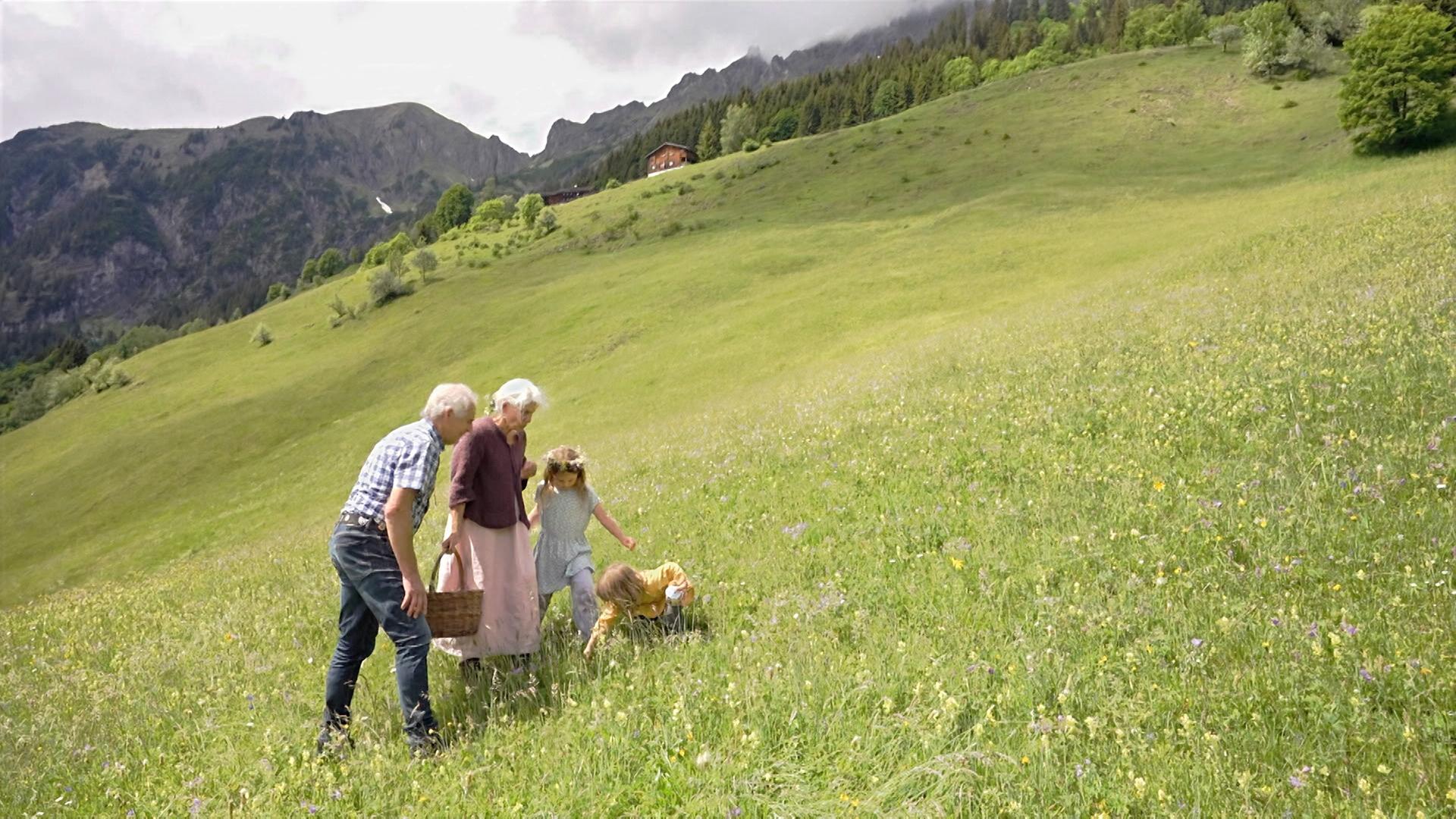 Im Bild sind drei Personen und ein Kind in einer grünen Wiese zu sehen. Sie gehen in Richtung einer Berglandschaft mit bewaldeten Hängen. Eine Person hält einen Korb.
