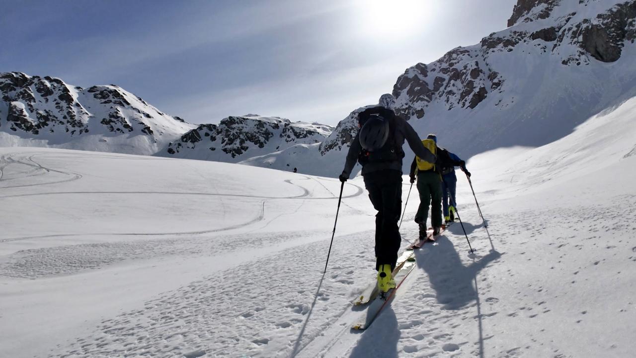 Im Bild sind vier Personen zu sehen, die auf Skiern einen schneebedeckten Hang hinaufsteigen. Im Hintergrund sind schneebedeckte Berge und ein blauer Himmel zu erkennen.