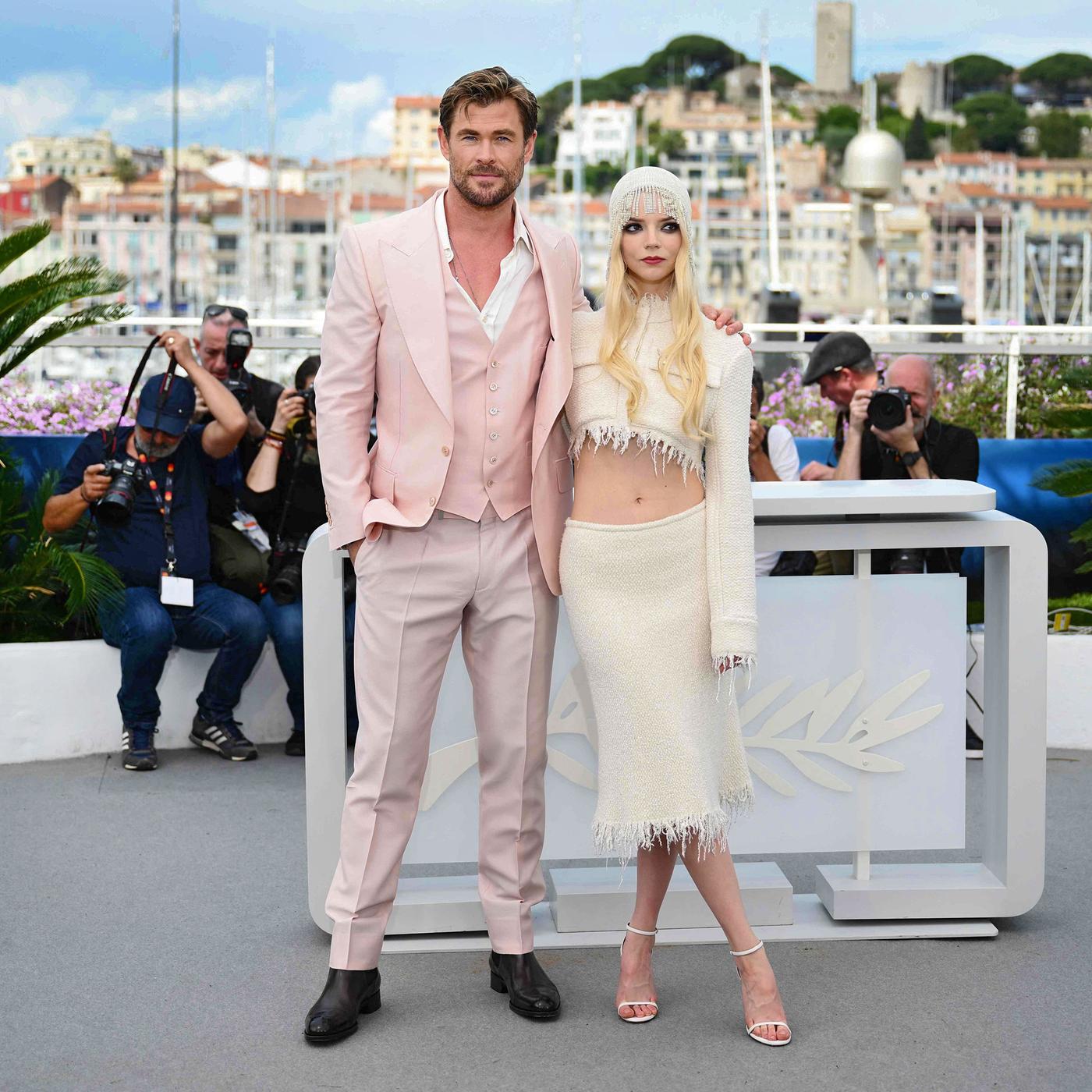 Ein Mann im lässig offenen rosa Jackett mit Weste und eine Frau in einem bauchfreien Kostüm posieren vor einem Pult mit Palmensymbol. Im Hintergrund sind Fotografen und ein Hafen zu sehen.