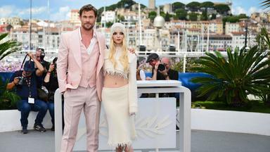 Ein Mann im lässig offenen rosa Jackett mit Weste und eine Frau in einem bauchfreien Kostüm posieren vor einem Pult mit Palmensymbol. Im Hintergrund sind Fotografen und ein Hafen zu sehen.